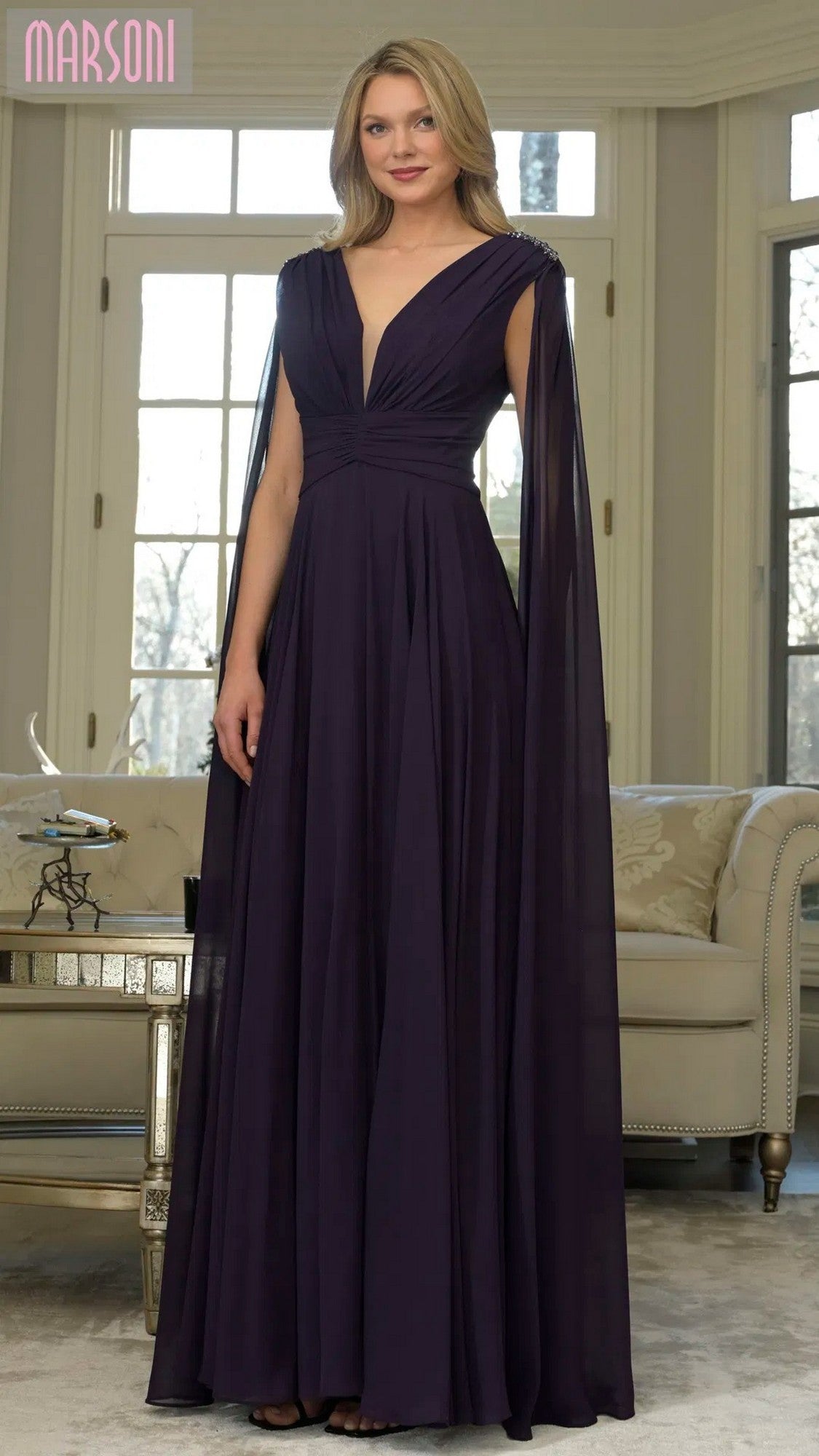 A woman stands indoors in front of large windows and a cream sofa, wearing the Marsoni MV1349 long formal dress in aubergine with a ruched bodice and long cape sleeves.
